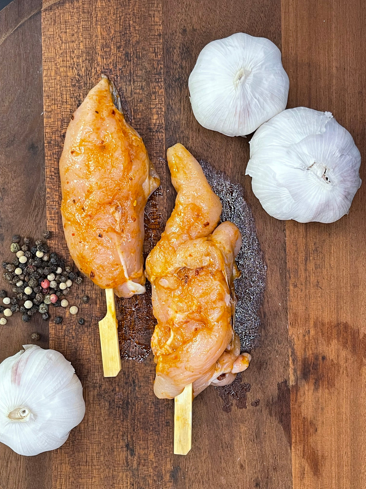 Two skewered marinated chicken pieces on a wooden cutting board with garlic and peppercorns.