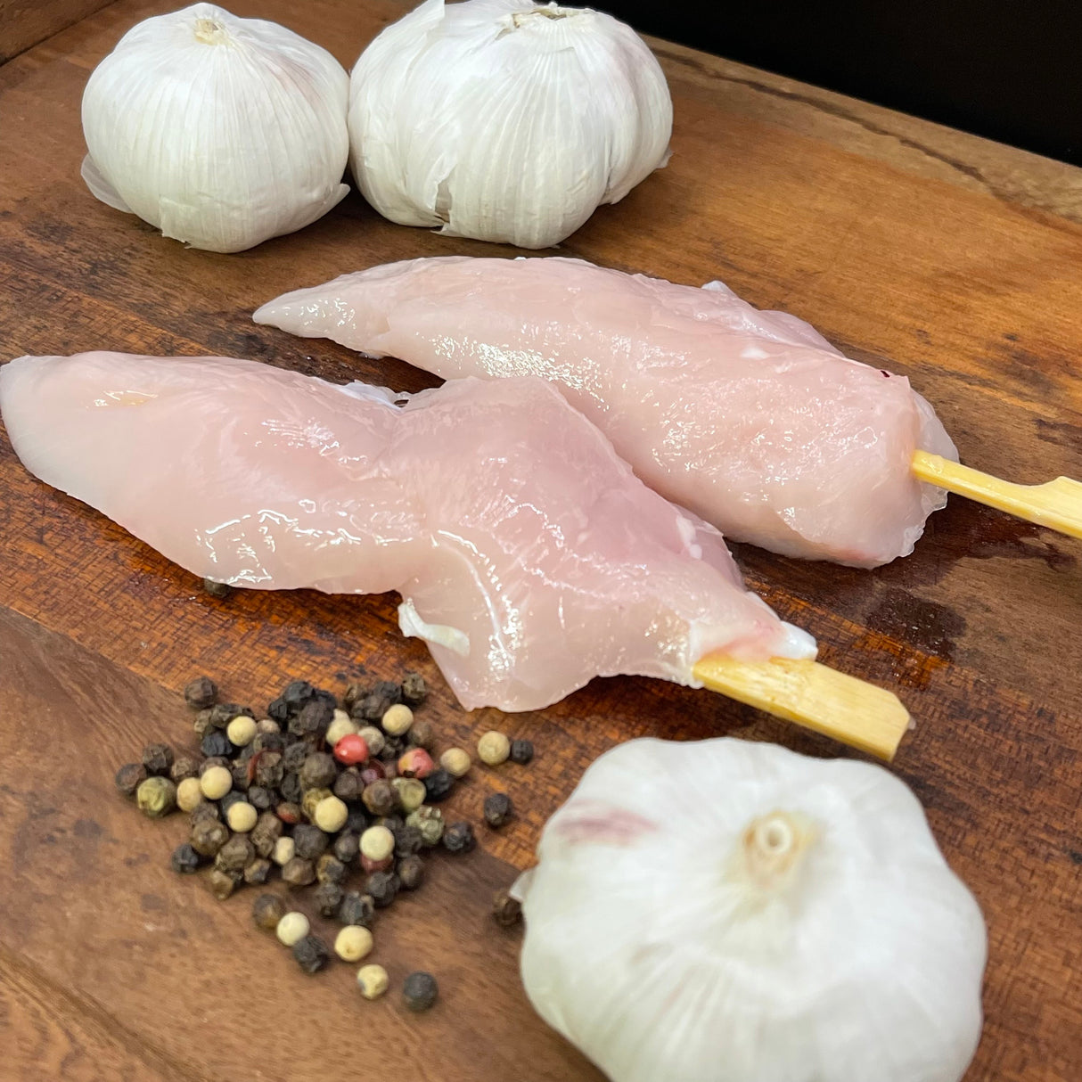 Raw chicken skewers on a wooden board with garlic and peppercorns.