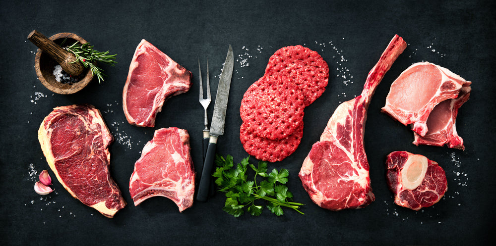 Assortment of raw cuts of meat, dry aged beef steaks and hamburger patties for grilling with seasoning and utensils on dark rustic board
