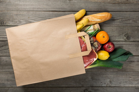 A paper bag overflowing with an assortment of red meat, fresh fruits and vegetables.