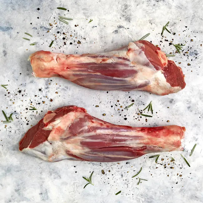 Two raw lamb legs on a textured surface with herbs and spices.